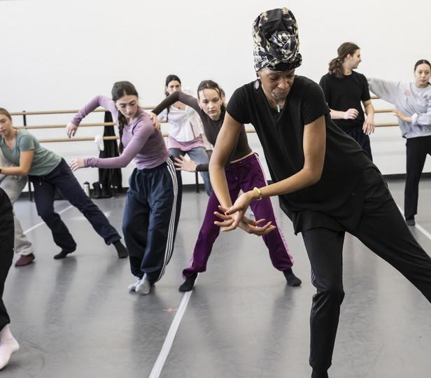 As part of a dance class, students practicing movements together. The central figure, a teacher, leads with dynamic energy, her body leaning forward in a guiding pose. She seems to be demonstrating a move, her hands outstretched as if to weave through the air, while her students, wearing various casual dance attire, mimic her stance with varying degrees of focus and precision. The mirrored wall reflects the concentrated ambiance of the room, creating an immersive environment of learning and movement.