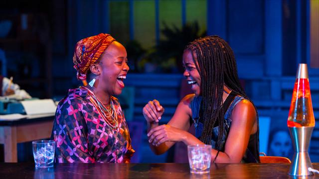 two actors laughing at a table on stage